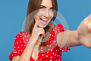 Cheerful young woman with bright smile in red casual dress looking to camera showing her white teeth over blue