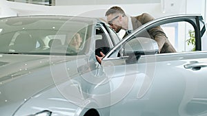 Cheerful young woman checking car interior in showroom talking to salesman