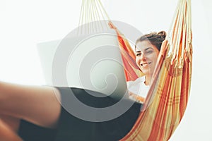 Cheerful young woman chilling in hammock with laptop. Freelancer and her workspace