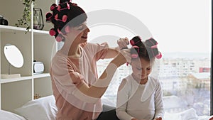 Cheerful young woman curls her hair while sitting on the bed near the big window