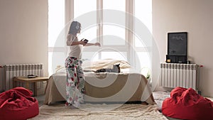 Cheerful young woman dancing in pajamas at home having fun listening to music wearing headphones enjoying weekend