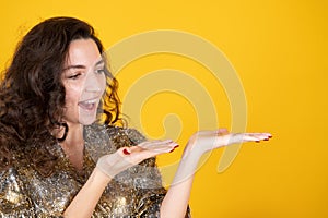 Cheerful young woman demonstrating something on her empty palm and pointing on it, yellow studio background