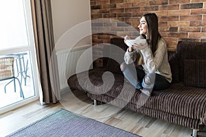 Cheerful young woman eating healthy breakfast while sitting on a couch at home