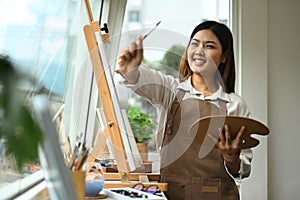 Cheerful young woman enjoying creative leisure activities, painting picture in bright modern art studio