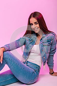 Cheerful young woman fashion model with beautiful smile in fashionable blue jeans wear sits and charmingly smiling near pink