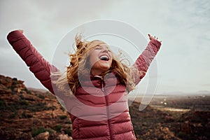 Cheerful young woman with hands raised and outstretched mountaineer with hair flying in wind enjoying fresh breeze -