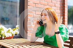 Cheerful young woman having conversation on mobile phone sitting at table in outdoor cafe terrace in sunny summer day.