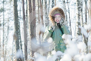 Cheerful young woman holding woolly mittens near her head in winter forest outdoors