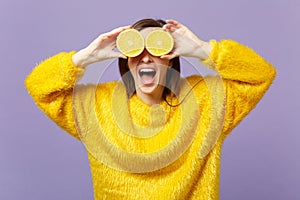 Cheerful young woman keeping mouth open, covering eyes with halfs of fresh ripe orange fruit isolated on violet pastel