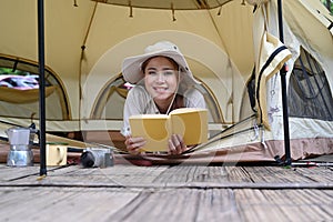 Cheerful young woman lying in her camping tent and reading a book. Travel, camping and vacation concept