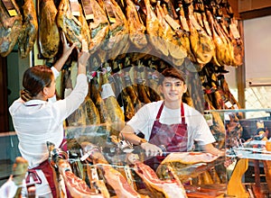 Cheerful young woman and man selling ham in jamoneria