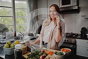 Cheerful young woman in modern kitchen preparing fresh vegetables salad and eating a carrot smiling at the camera
