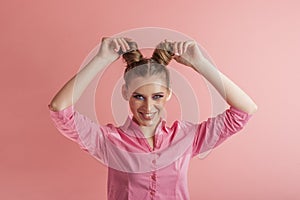 Cheerful young woman in pink shirt adjusts her hair. Girl with two bun hairstyles on pink background