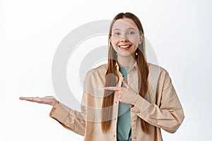 Cheerful young woman pointing finger at open hand, showing product, demonstrate promo item on palm against copyspace