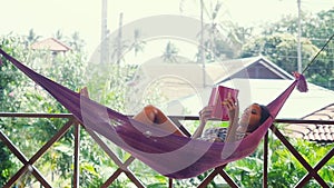 Cheerful young woman relaxing in a peaceful garden during summer holiday and reading a book in a swinging hammock. Slow