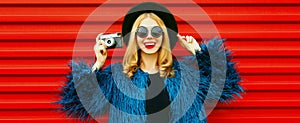 Cheerful young woman with retro camera wearing blue faux fur coat, round hat over red