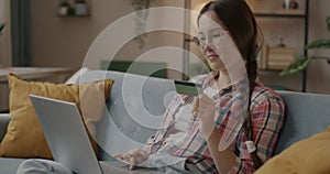 Cheerful young woman shopping in online store using laptop paying with credit card indoors at home in evening