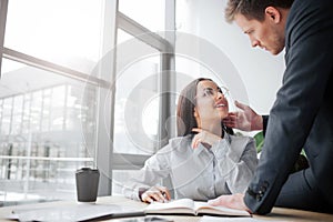 Cheerful young woman sit at table and smile to her boss. She look at him. Guy is sexually excited. He touches her face.