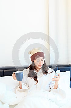 Cheerful young woman sitting in bed texting on her smartphone and drinking coffee at home in bedroom