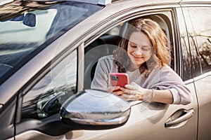 Cheerful young woman sitting in a car in the driver& x27;s seat looking into a smartphone, paying for parking and