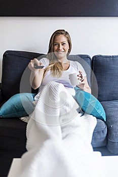 Cheerful young woman sitting on a couch at home while using a remote tv controller