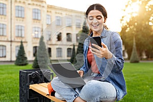 Cheerful young woman student using laptop and phone outdoors