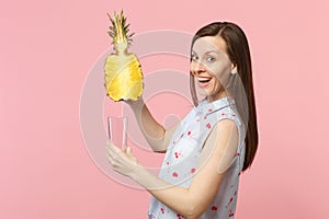 Cheerful young woman in summer clothes holding half of fresh ripe pineapple fruit glass cup isolated on pink pastel wall