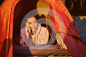 Cheerful young woman talking on mobile phone in camp tent