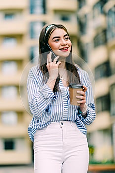 Cheerful young woman talking phone and holding a paper cup on the street