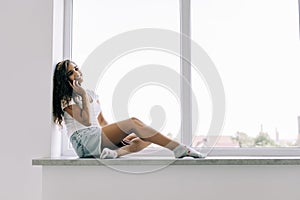 Cheerful young woman talking phone and sitting on windowsill at home