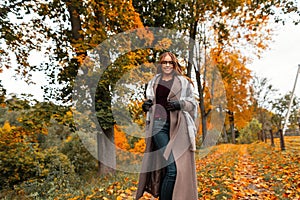 Cheerful young woman in trendy warm autumn clothes runs along a path in a park with trees with yellow-orange leaves. Cheerful girl