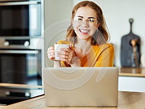 cheerful young woman uses a laptop at home in kitchen with cup of morning coffee