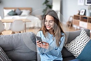 Cheerful young woman using mobile phone while sitting on a couch at home