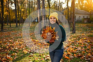 Cheerful young woman walking in autumn park at beautiful sunset and collecting a dry bouquet of fallen colorful leaves. Sun Rays