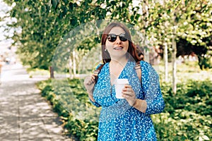 Cheerful young woman wearing blue dress and sunglasess walking outdoors, happy girl holding takeaway coffee cup on city
