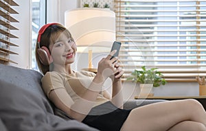 Cheerful young woman wearing headphones and using smart phone in cozy living room