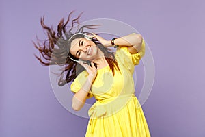 Cheerful young woman in yellow dress with fluttering hair listen music putting hands on headphones  on pastel