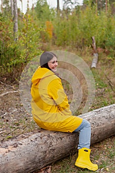 cheerful young woman in a yellow raincoat and yellow shoes is sitting on a fallen log.