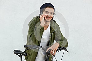 Cheerful youngster with little beard, has pleasant talk with girlfriend via smart phone, wears fashionable hat and anorak, leans a