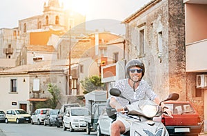 Cheerfully smiling man in helmet and sunglasses fast riding the moto scooter on the Sicilian old town streets with parked cars in