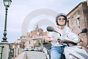 Cheerfully smiling woman in helmet and sunglasses fast riding the moto scooter on the Sicilian old town streets in the Forza d`