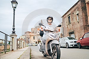 Cheerfully smiling woman in helmet and sunglasses fast riding the moto scooter on the Sicilian old town streets in the Forza d`