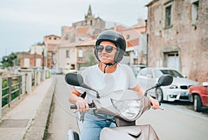 Cheerfully smiling woman in helmet and sunglasses fast riding the motor scooter on the Sicilian old town streets in the Forza d`