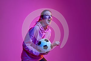 Cheering the team up. Portrait of young emotive girl posing with football ball background in neon light. Concept of