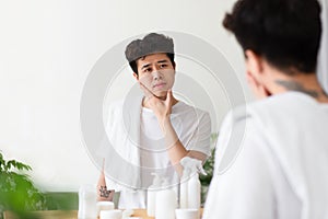 Cheerless asian young man with tattoos in white t-shirt, with towel, touches hand to cheek, checks stubble