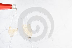 Cheers background with flow of pouring sparkling champagne from red neck bottle to two glasses on light white marble backdrop.