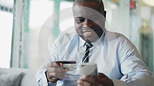 Cheery African American businessman in formal clothes paying online bill keeping credit card and smartphone in his hands