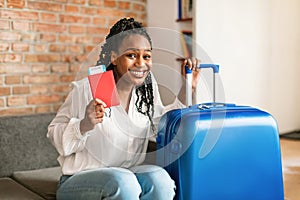 Cheery african american woman sitting next to suitcase, holding passport and plane tickets, getting ready for trip