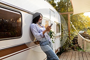 Cheery Asian lady leaning on motorhome, using cellphone on autumn camping trip in countryside