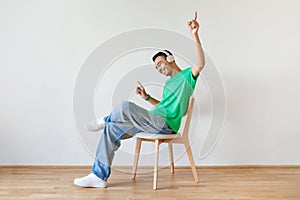 Cheery guy in headphones listening to music and dancing while sitting on chair against white studio wall, free space